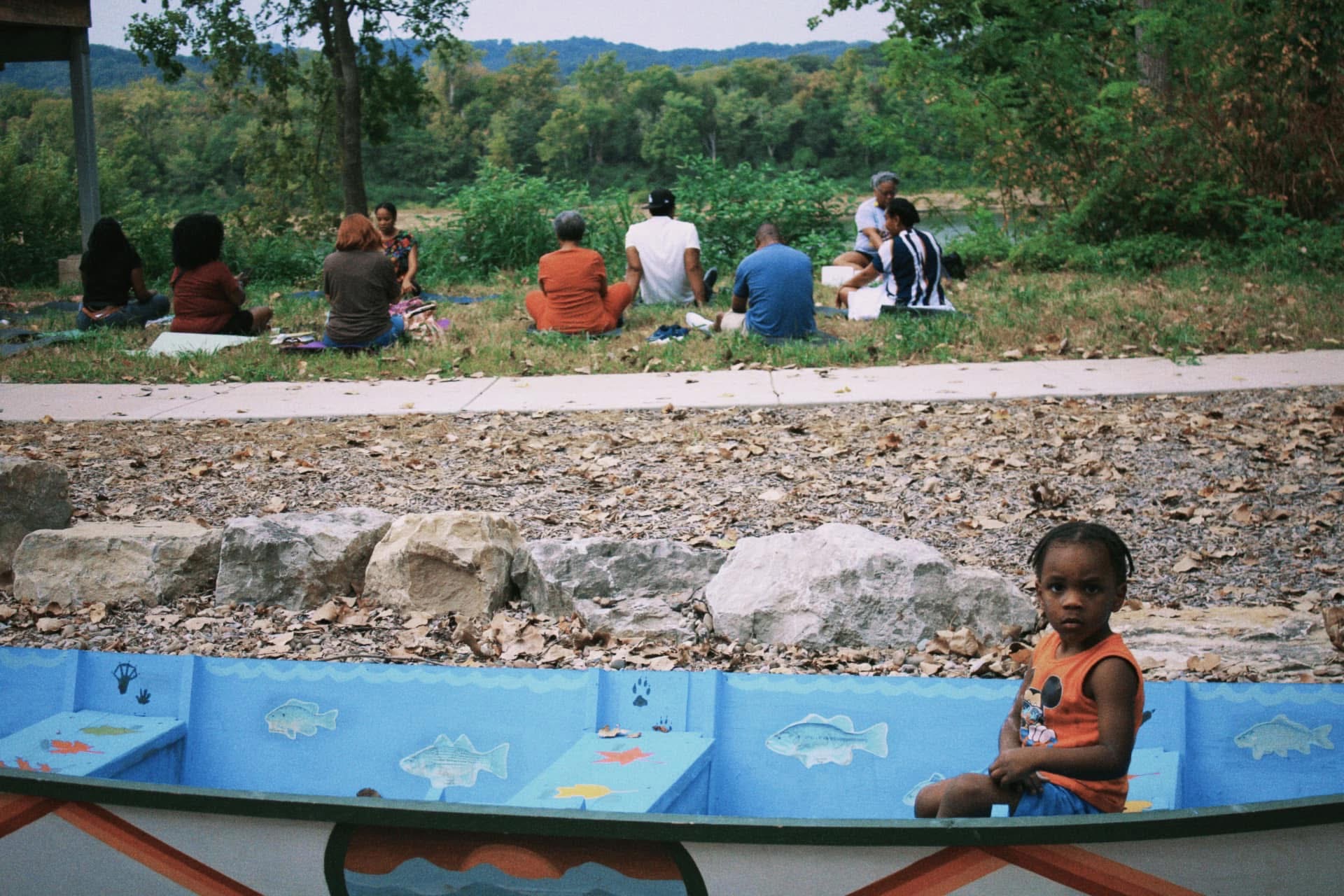 A child resting on a painted canoe bench while elders meditate by the river