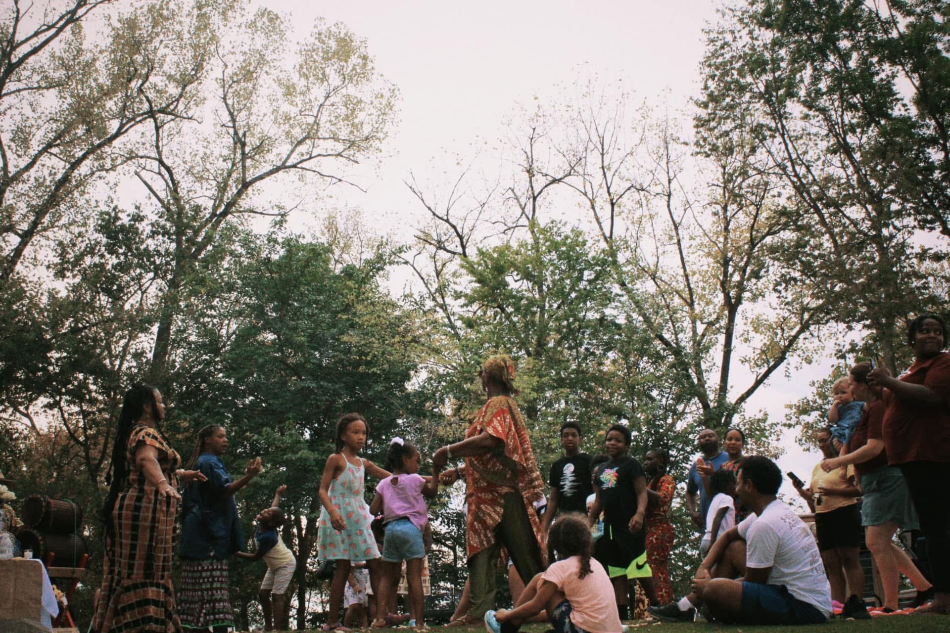 Kids and elders dancing together in a circle under tall trees
